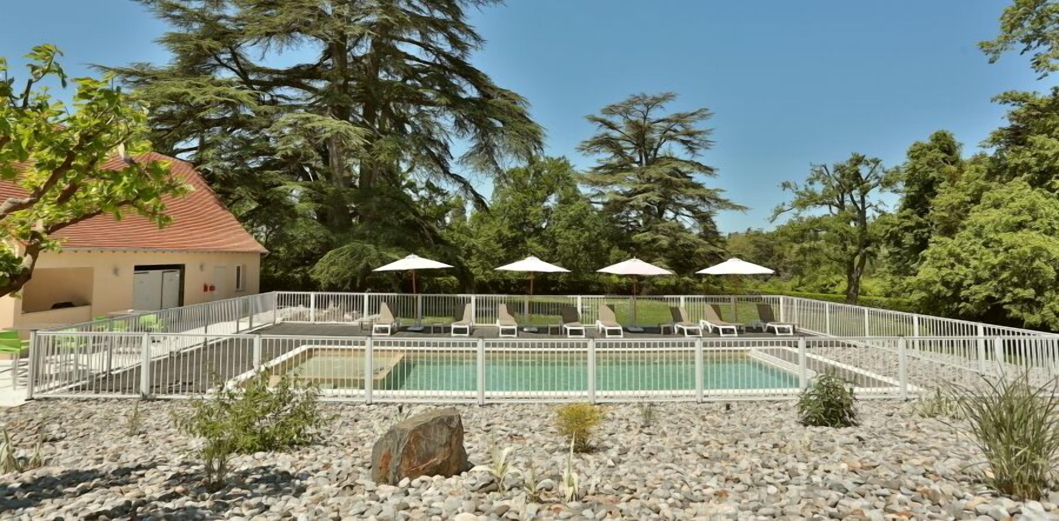 Zona de piscina con sillones de descanso y sombrillas, rodeada por una valla blanca y un edificio con techo rojo.