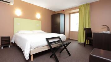 Una habitación de hotel con cama, escritorio, silla, armario y ventana con cortinas.