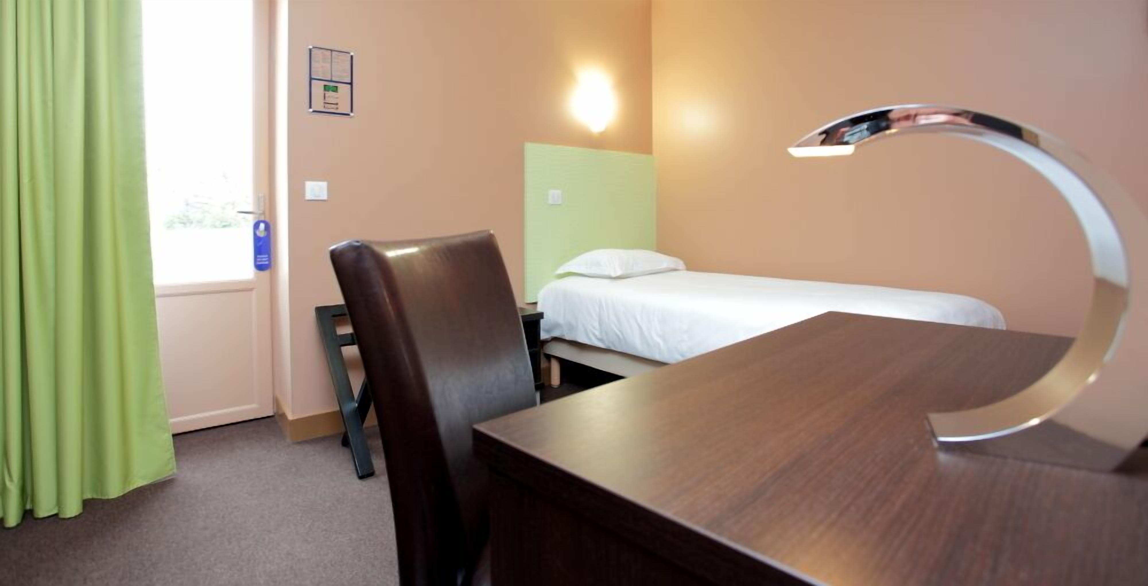Et hotelværelse med seng, skrivebord, stol, skrivebordslampe og et vindue med gardiner.
