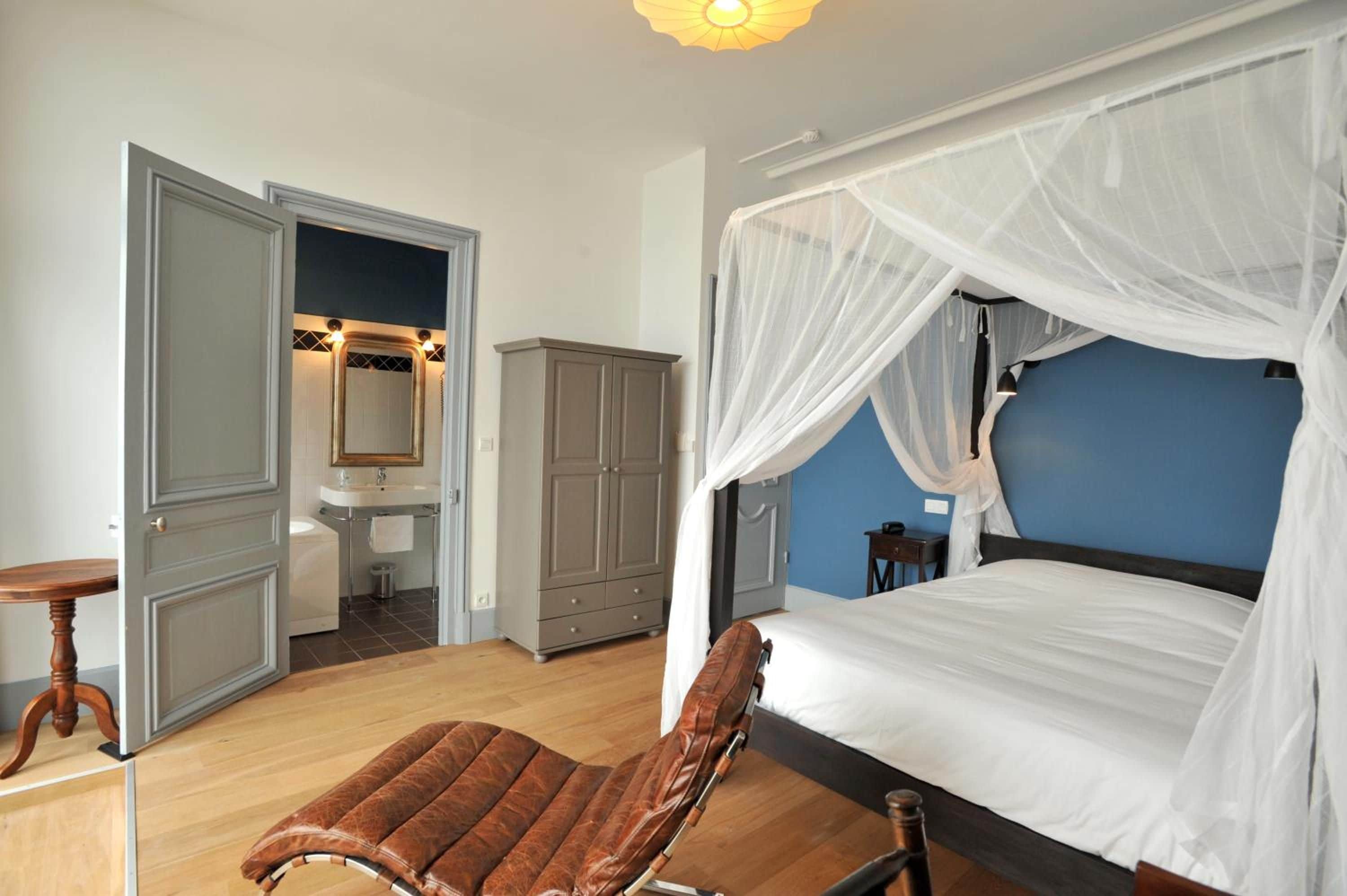 A bedroom with a four-poster bed, a wooden bench, a wardrobe, and a bathroom with a mirror and sink.