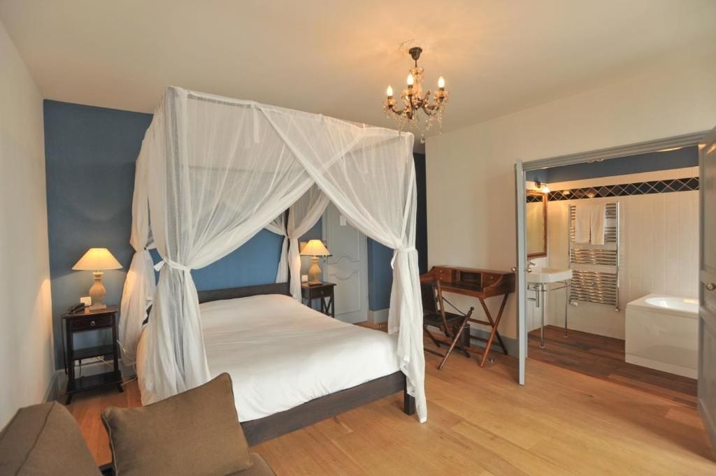 A bedroom with a four-poster bed, bedside lamps, a desk, and a view into the bathroom.