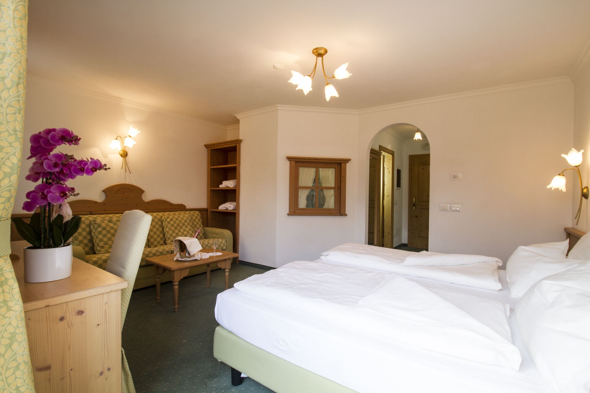 A hotel room with a large bed, a small desk, a chair, a wooden wardrobe, and a potted orchid.