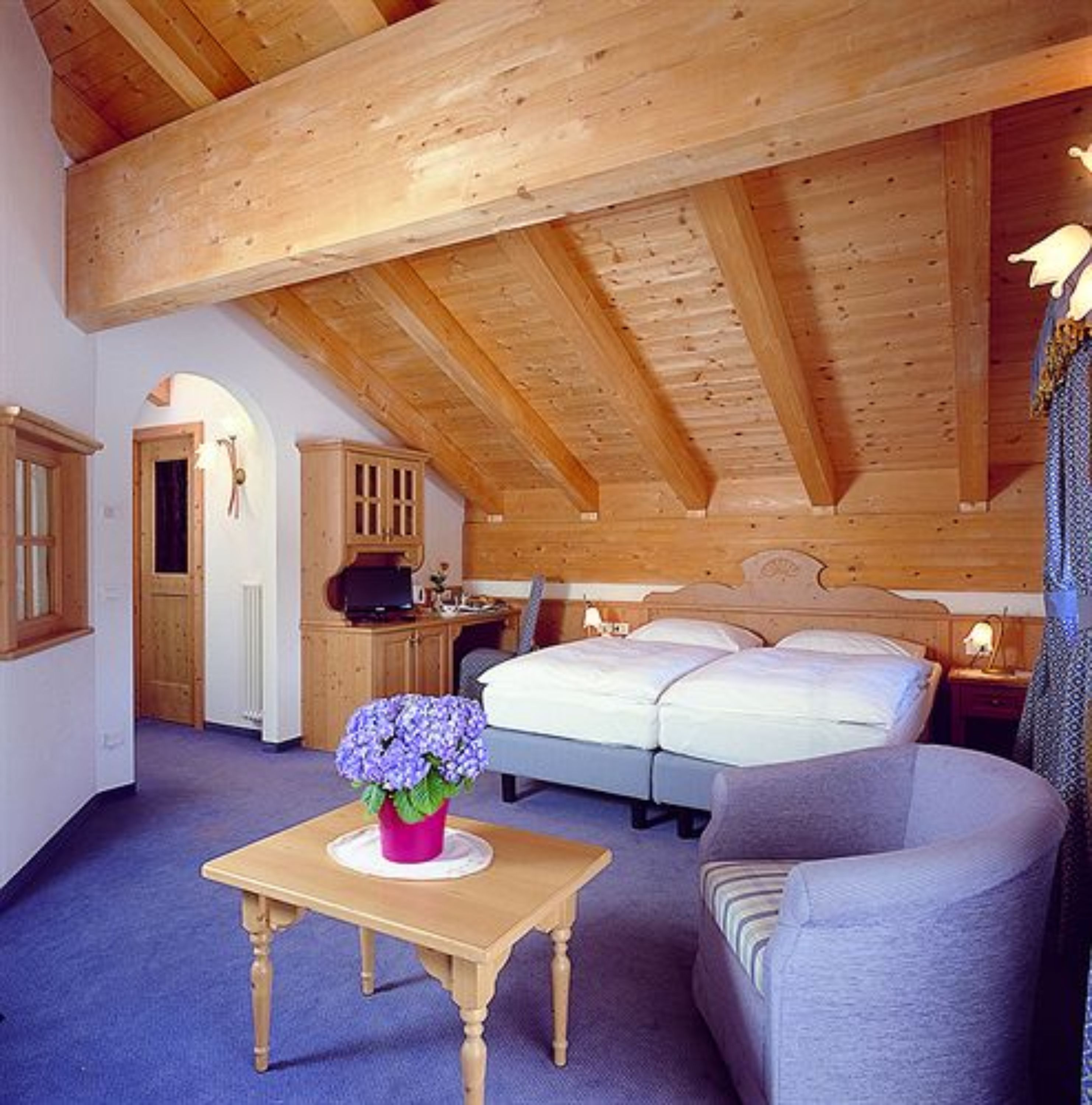 Een slaapkamer met een houten plafond, een bed, een nachtkastje, een dressoir en een kleine tafel met een bloemenvaas.