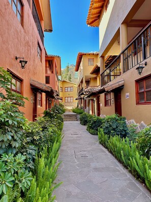 Un estrecho callejón bordeado por edificios con balcones, escalones de piedra que conducen a una puerta y abundante vegetación.