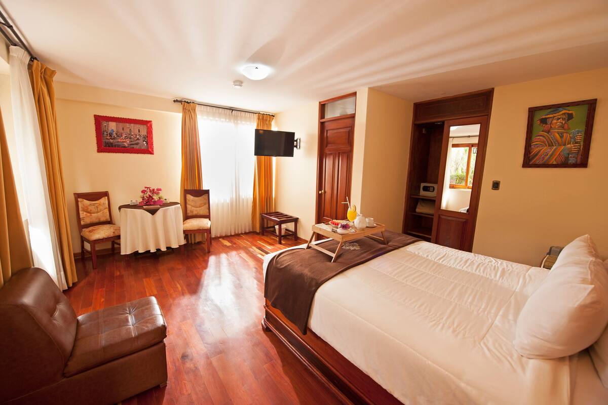 A hotel room with a large bed, a wooden floor, a TV mounted on the wall, and a small table with a chair.