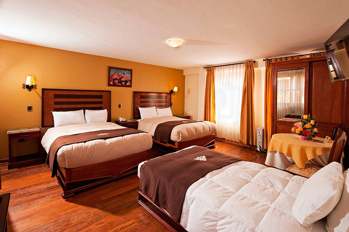 A hotel room with two beds, wooden headboards, a desk with a chair, a small table with a vase of flowers, and a painting on the wall.