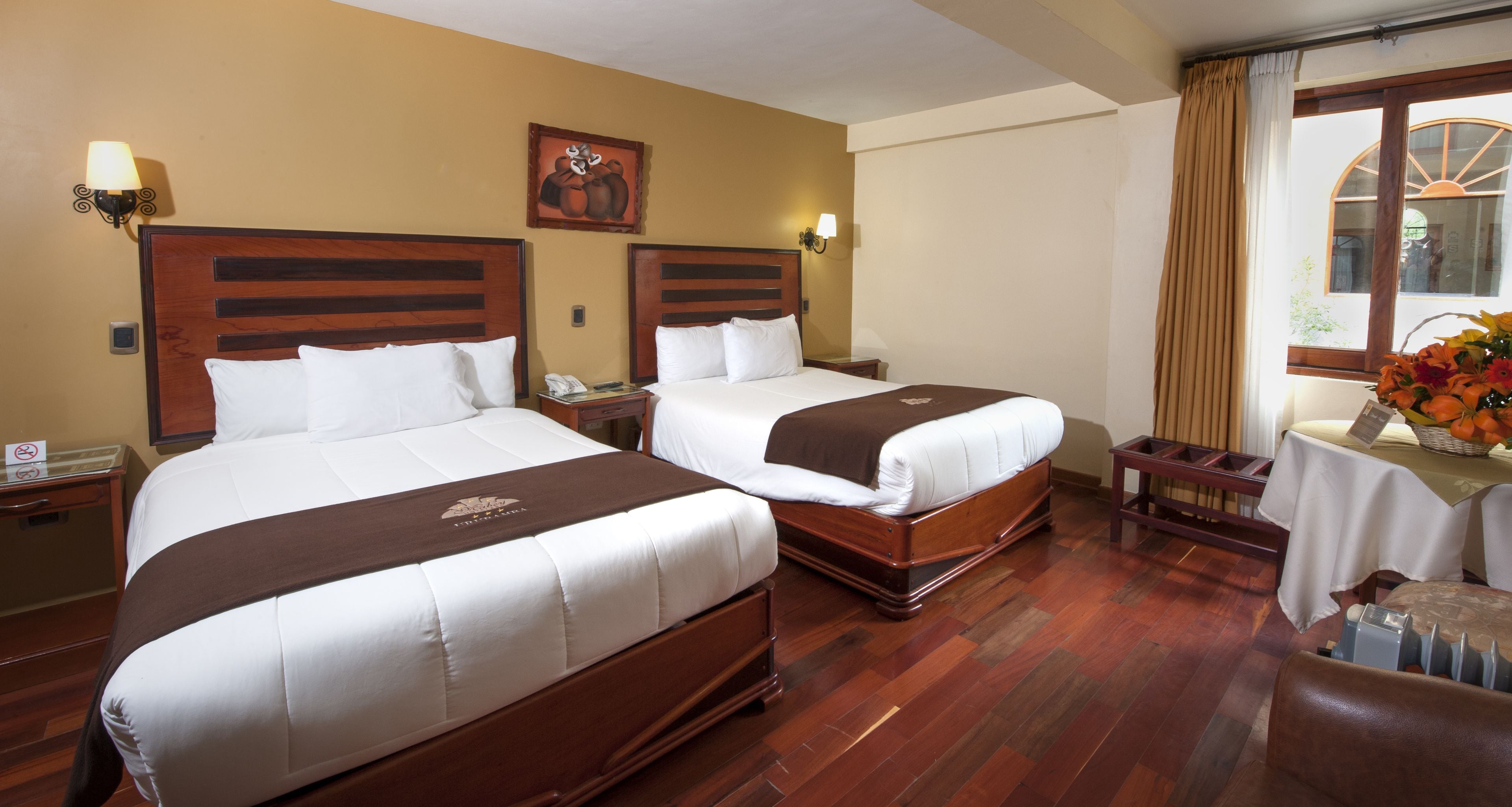 A hotel room with two beds, wooden headboards, a small table with a vase of flowers, and a window with curtains.