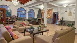 A spacious living room with beige sofas, a glass coffee table, and a fireplace. There are large windows and a chandelier. - Hotel Mabey Urubamba (Urubamba)