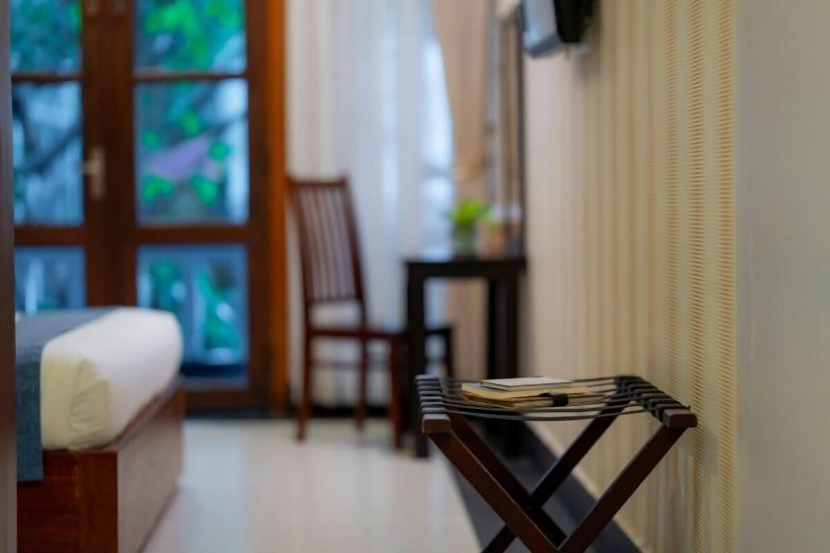 A room with a bed, a wooden chair, a small table with a book, and a window with a view of greenery.