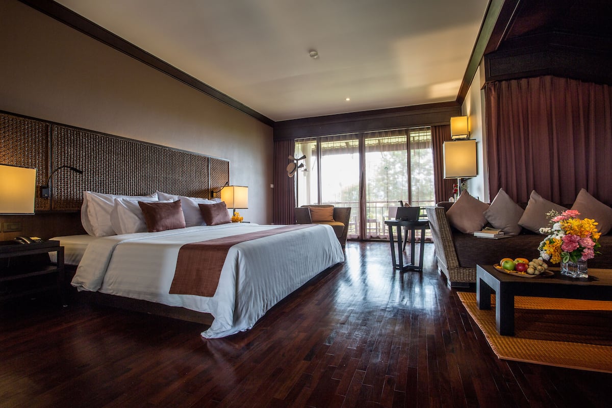 A hotel room with a large bed, a sofa, a dining table with fruit, and a view of the outdoors.