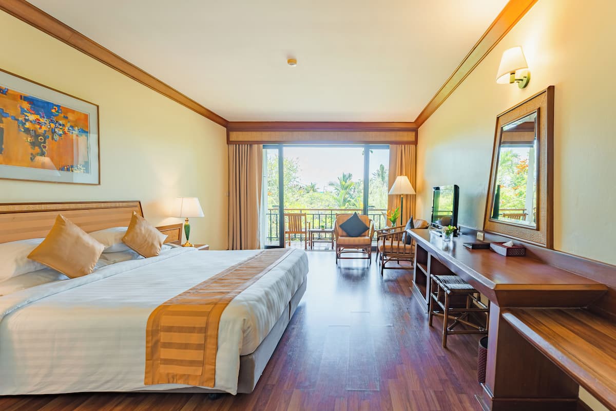A hotel room with a large bed, a desk with a computer, a chair, and a balcony with a view of trees.