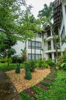 Un edificio de varios pisos con un jardín que incluye un camino de piedra, plantas diversas y una palmera.