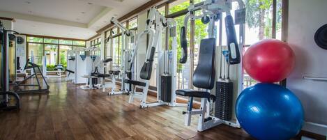 Uma academia com diversos equipamentos de exercício, janelas amplas e piso de madeira.
