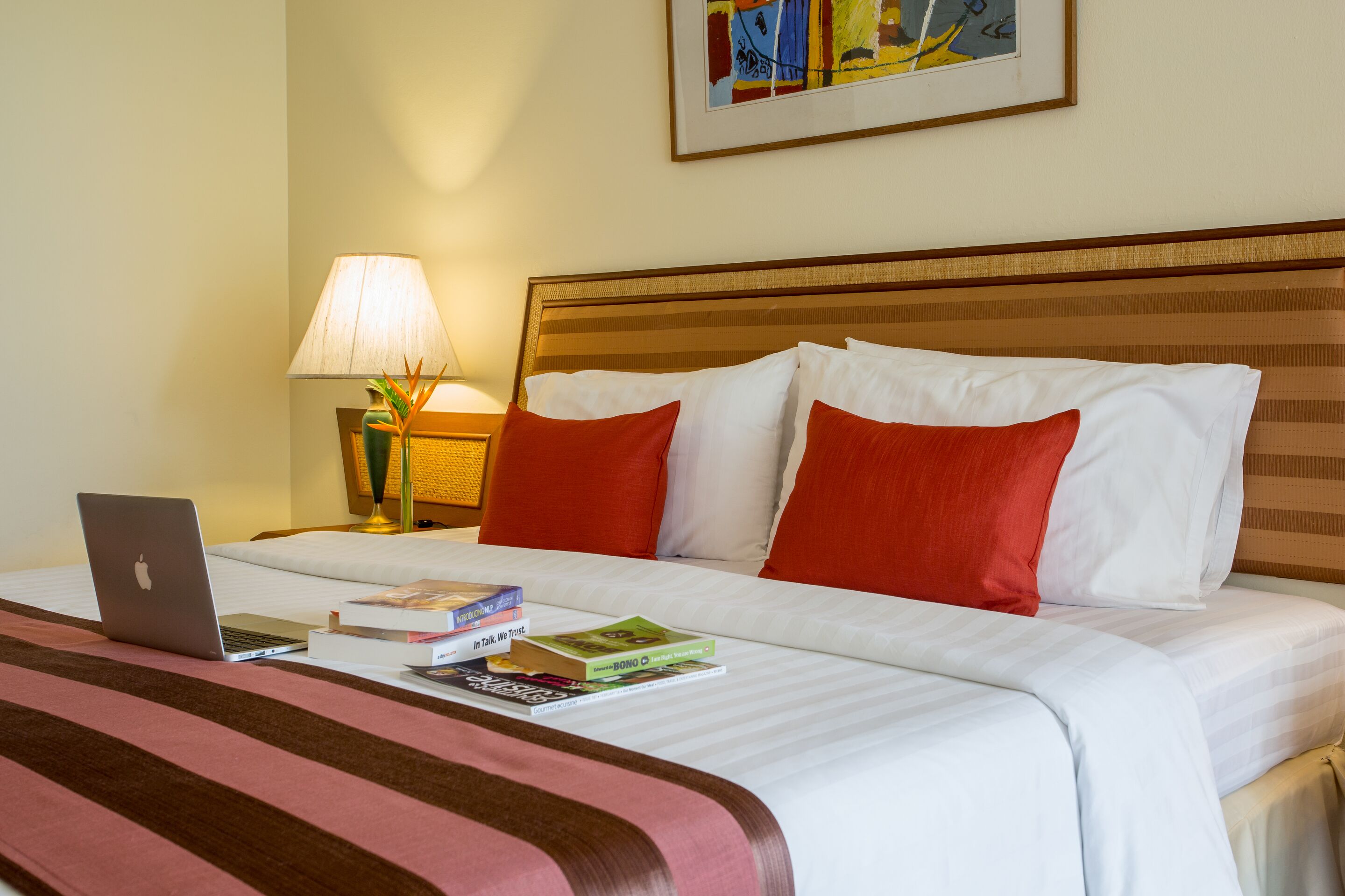 Una cama bien hecha con almohadas blancas y rojas, una computadora portátil y varios libros sobre la cama.
