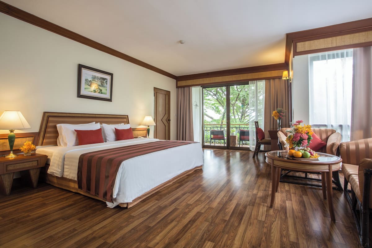 A hotel room with a large bed, a sitting area with a table and chairs, and a balcony with a view of trees.