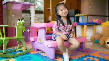 A child sitting on a pink bench in a play area with colorful mats and toys, including a rocking horse and a teddy bear.