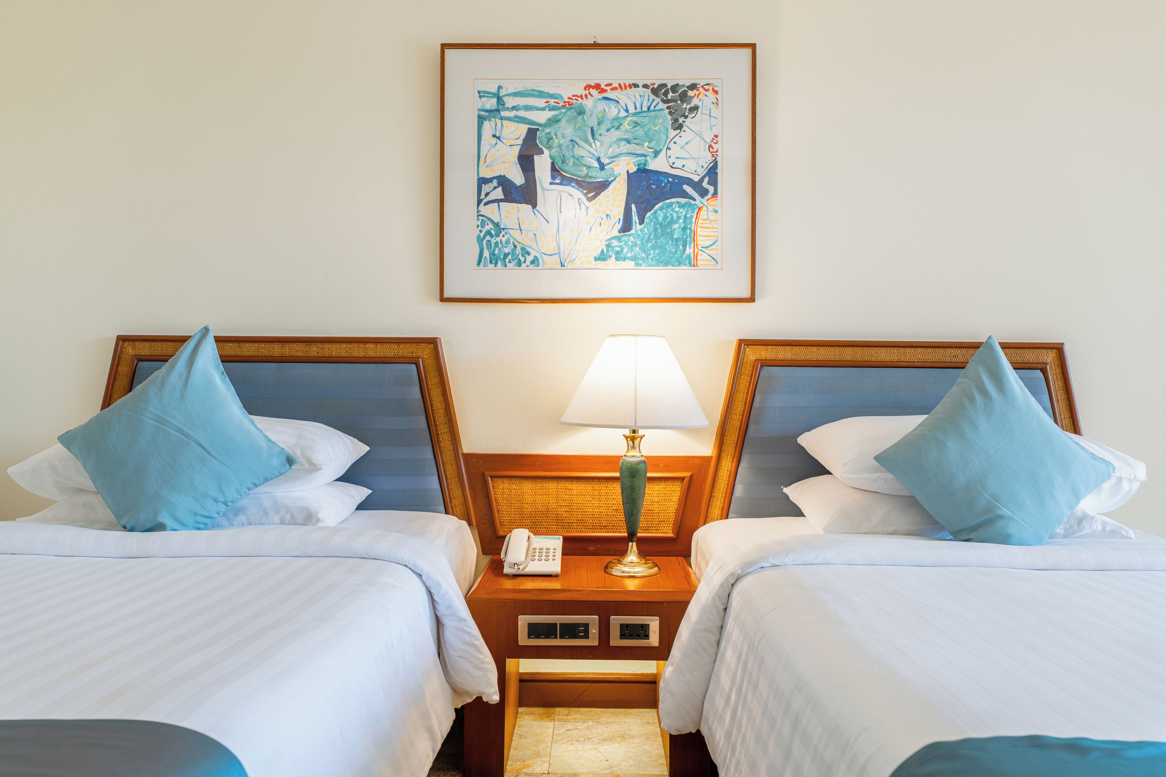 A hotel room with two beds, each with a blue pillow, a bedside table with a lamp, and a framed abstract painting on the wall.