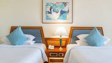 A hotel room with two beds, each with a blue pillow, a bedside table with a lamp, and a framed abstract painting on the wall.