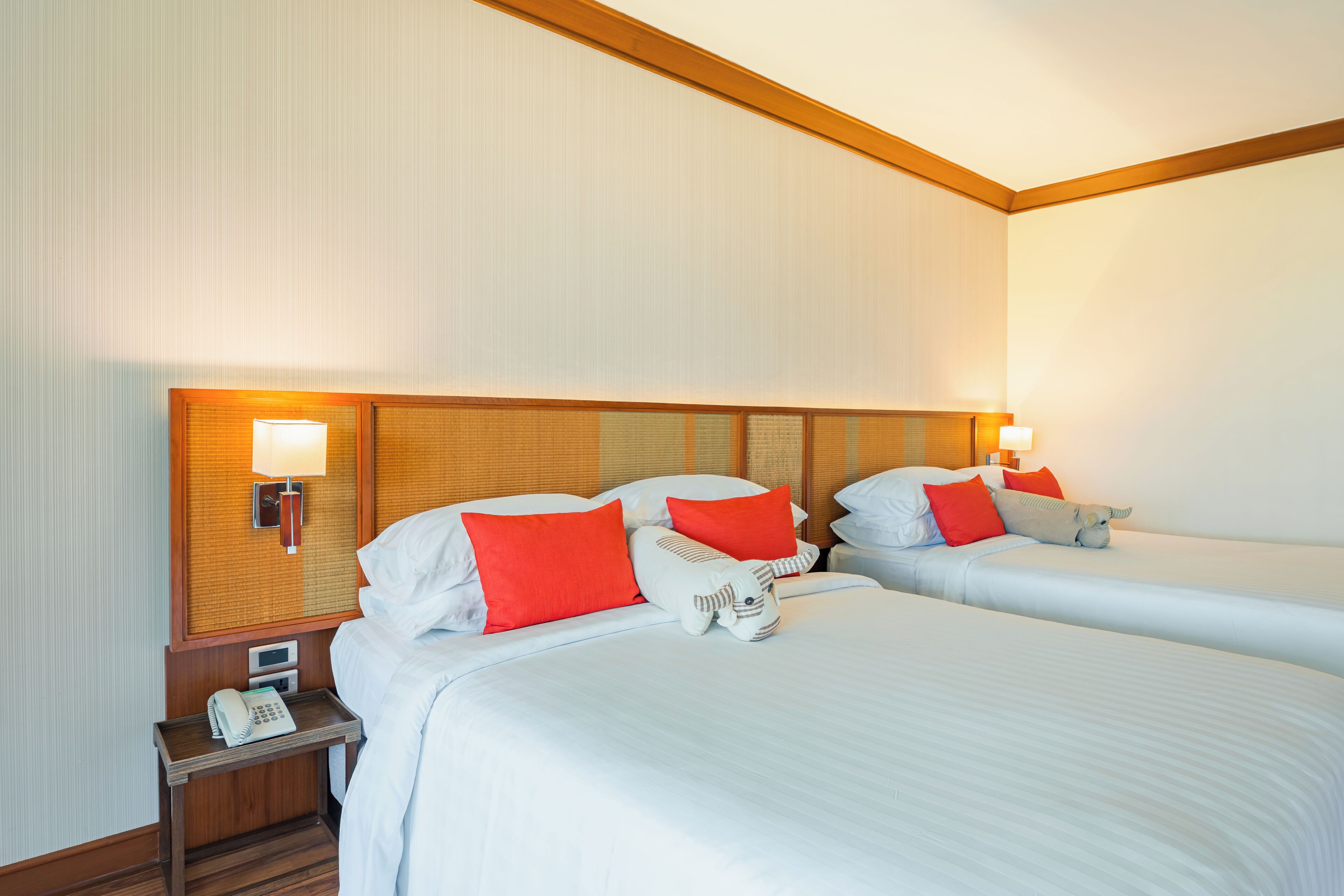 Habitación de hotel con dos camas, cada una con ropa de cama blanca y almohadas rojas. Hay lámparas de noche, una mesita de noche con un teléfono y un reloj en la pared.