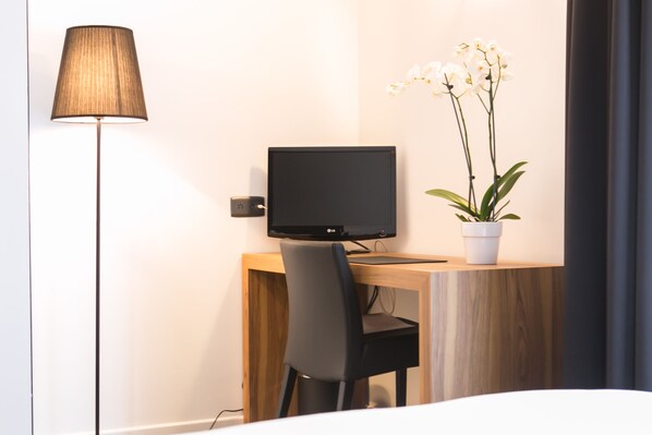 Una habitación de hotel moderna con un escritorio de madera, una silla negra, un televisor de pantalla plana, una lámpara y una orquídea en maceta.