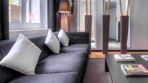 A modern living room with a dark grey sofa, white cushions, a lamp, and a magazine rack. - Saint James (Mons)