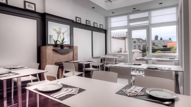 A modern dining area with white tables and chairs, framed artwork on the walls, and a view of buildings through large windows. - Saint James (Mons)