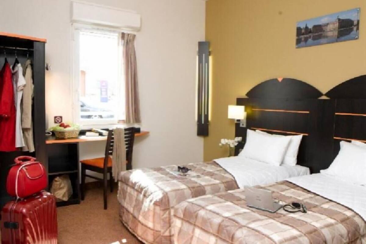 A hotel room with two beds, a desk with a chair, a red suitcase, and a window with curtains.