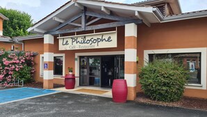 Un café-restaurant appelé «Le Philosophe», avec une entrée bien visible, un espace de stationnement bleu et des plantes en pot.
