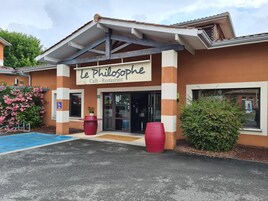 Un café y restaurante llamado "Le Philosophe" con entrada visible, zona de estacionamiento azul y plantas en macetas.