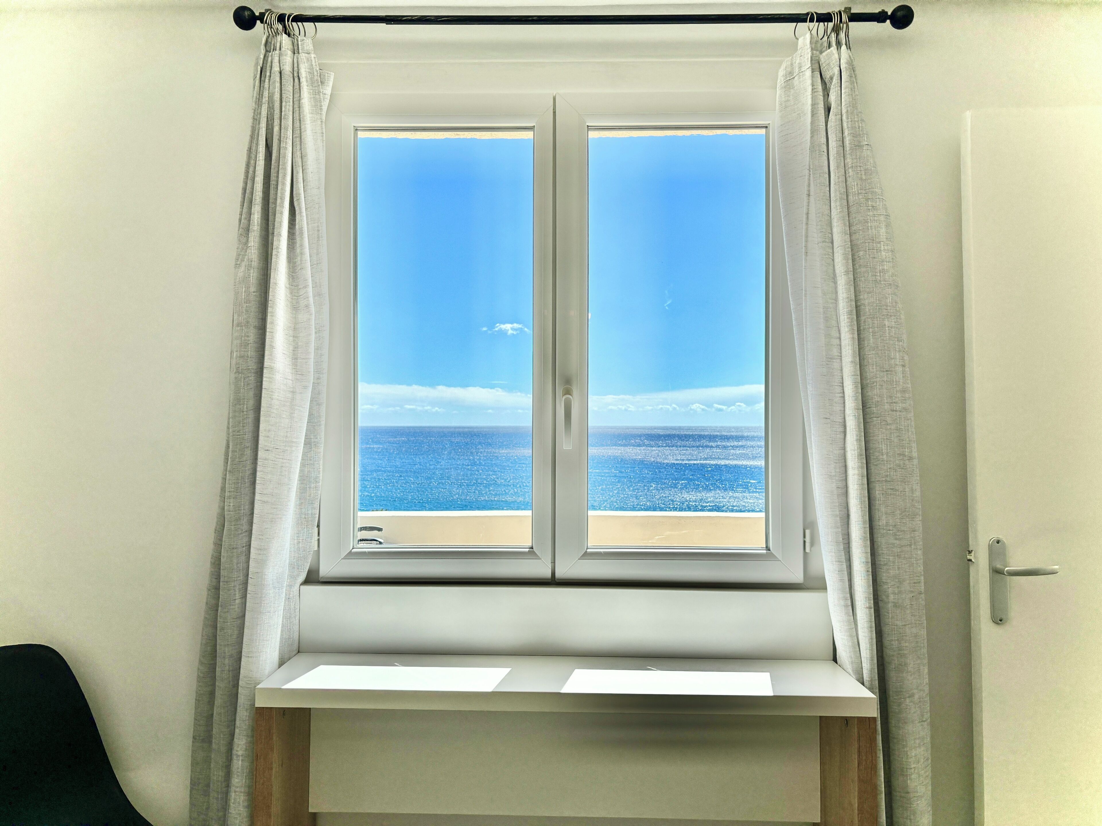 A room with a window overlooking a beach, a white door, and a black chair.