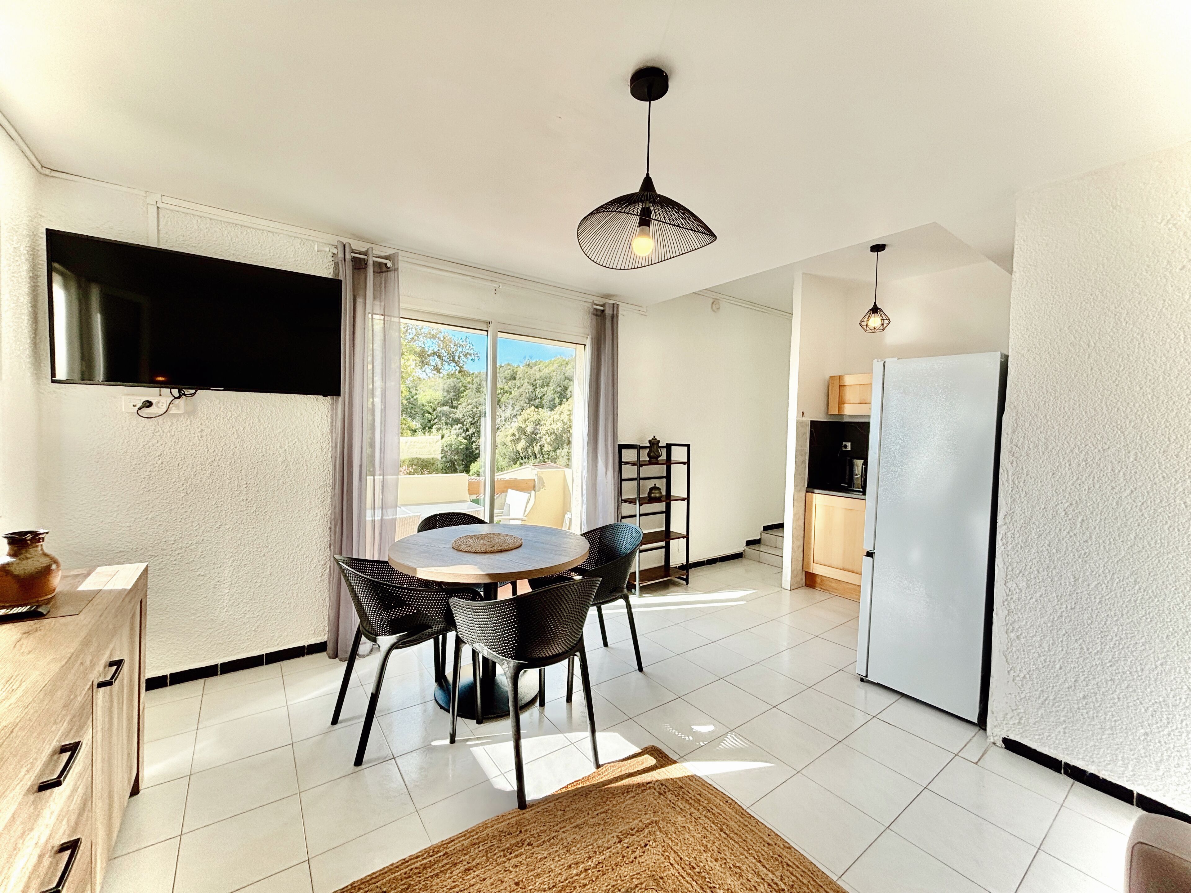 A compact kitchen with a dining area, a TV mounted on the wall, and a view of the outdoors through the sliding glass door.