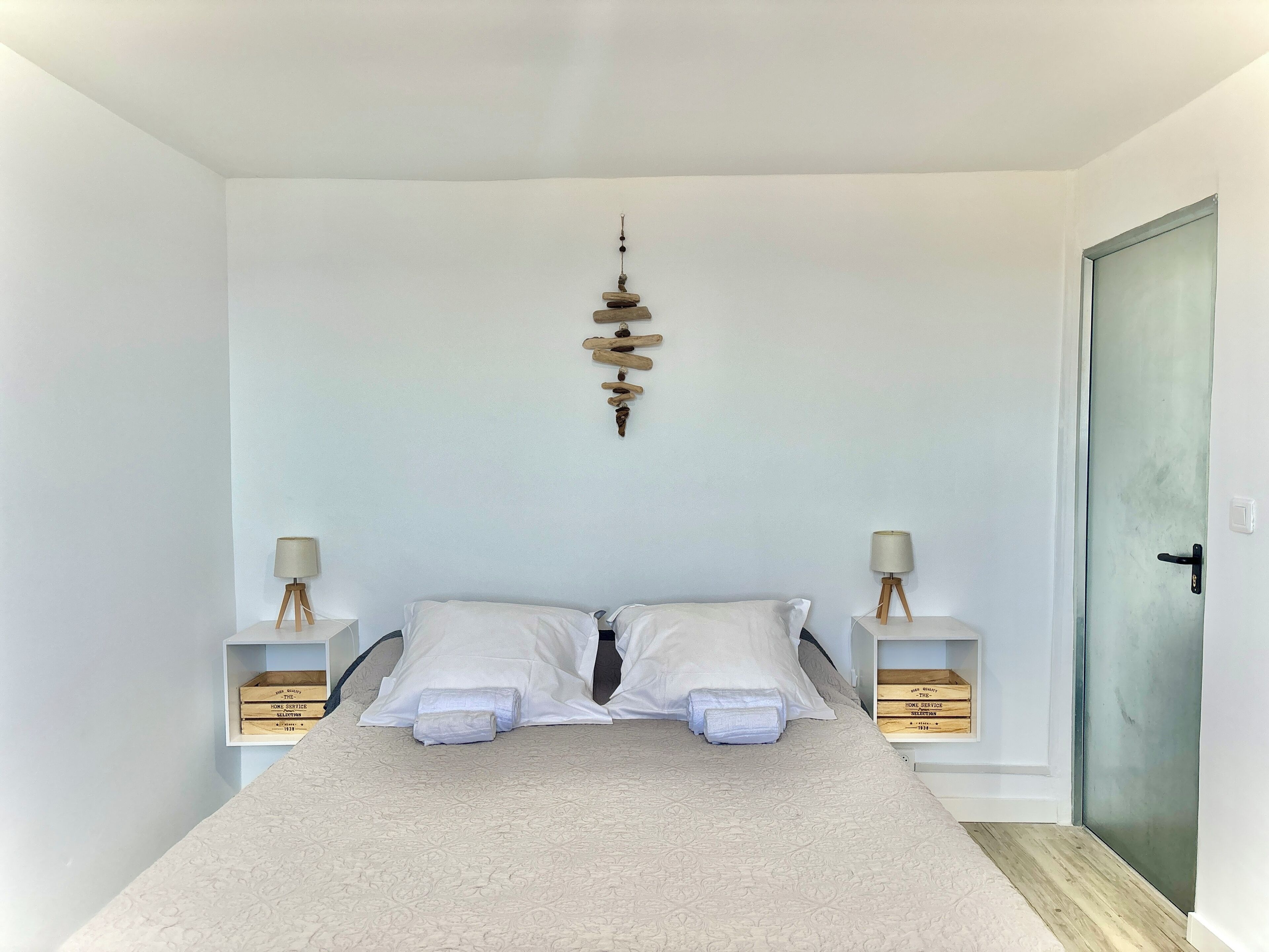 Une chambre à coucher bien rangée, avec un lit placé au centre, entre deux tables de chevet, chacune ornée d’une lampe et d’une boîte en bois.
