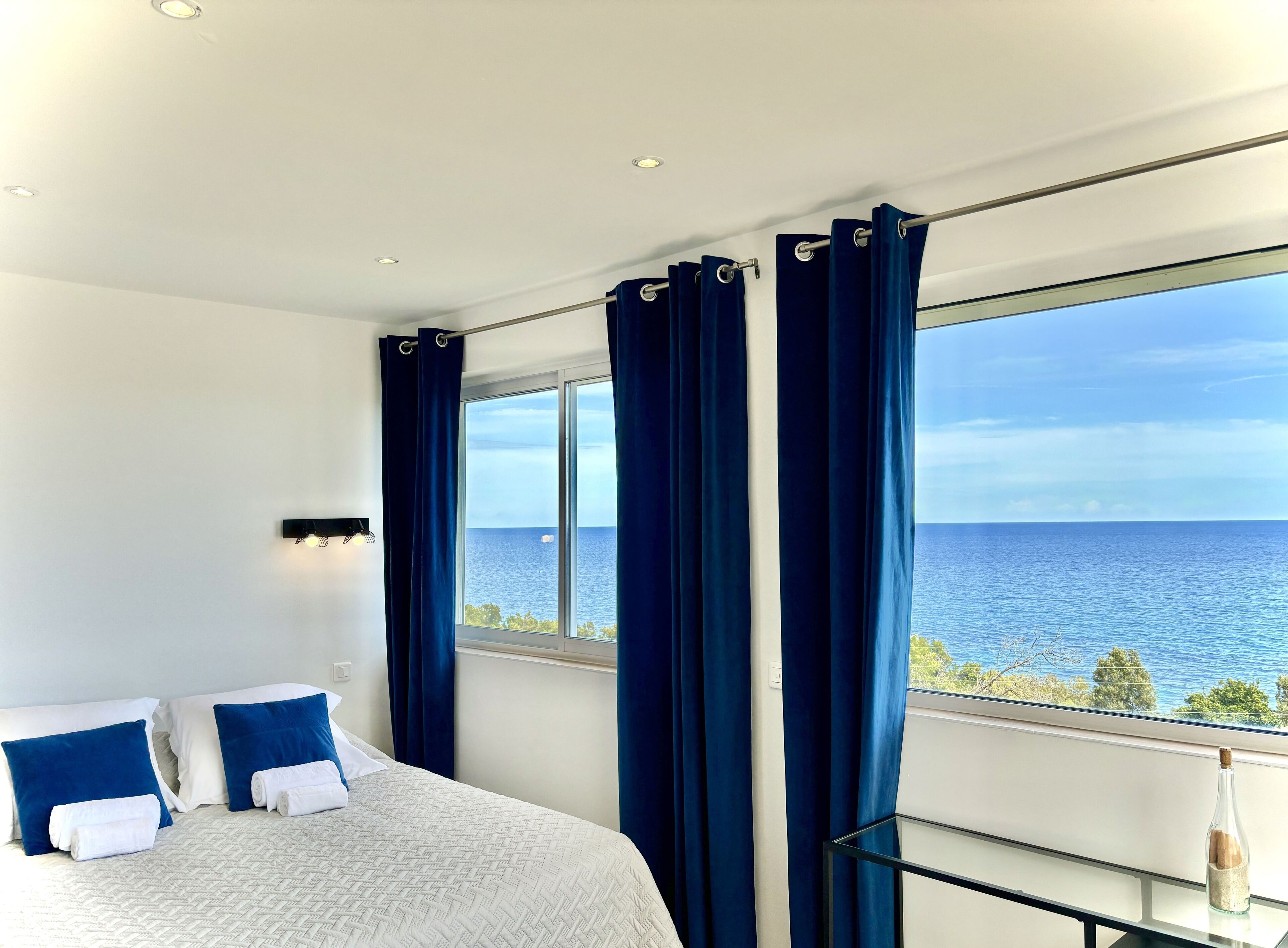 Une chambre avec une grande fenêtre, un lit avec une literie blanche et bleue, et une vue sur la mer.