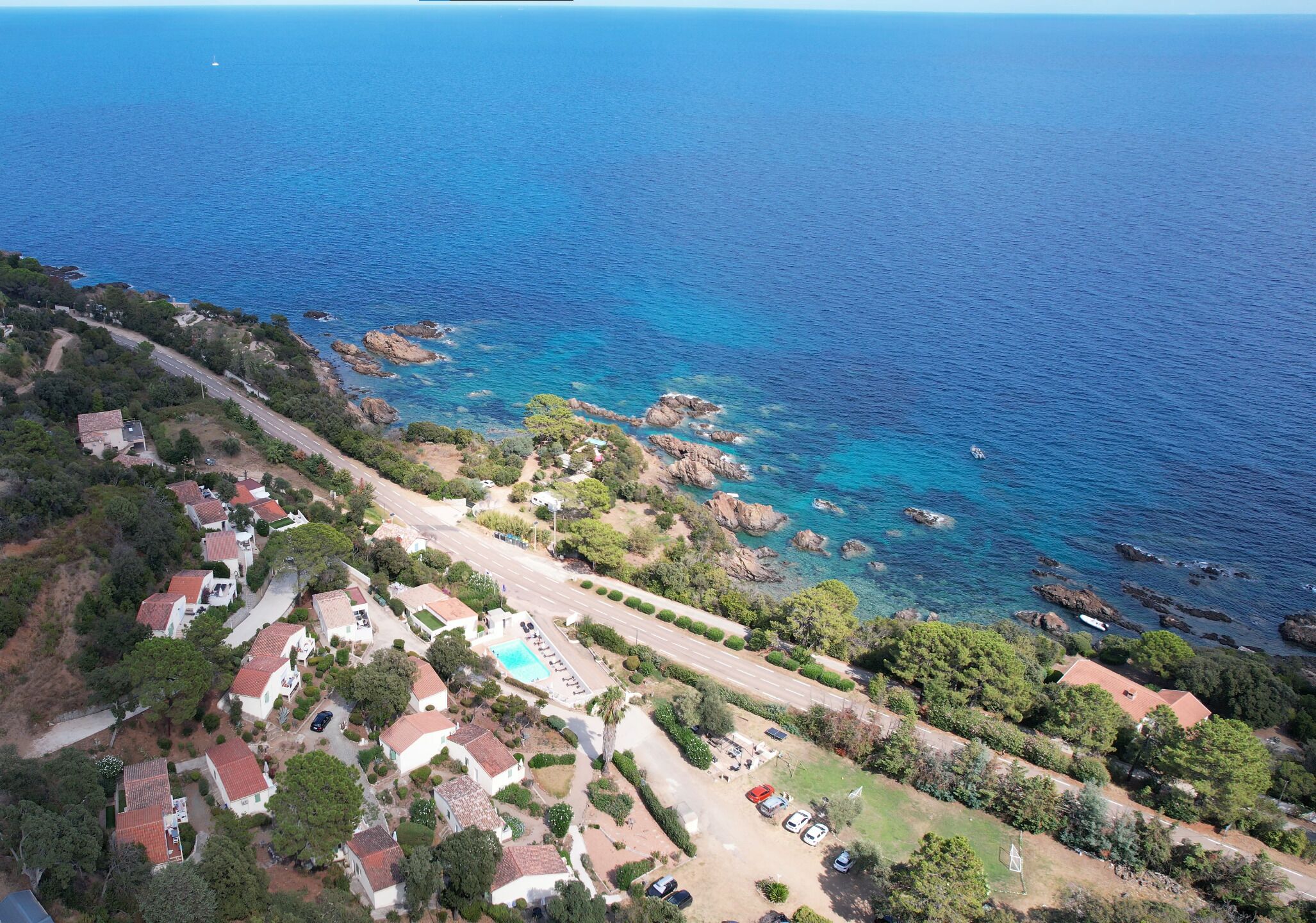 An aerial view of a coastal residential area with houses, a swimming pool, and a road leading to the sea.