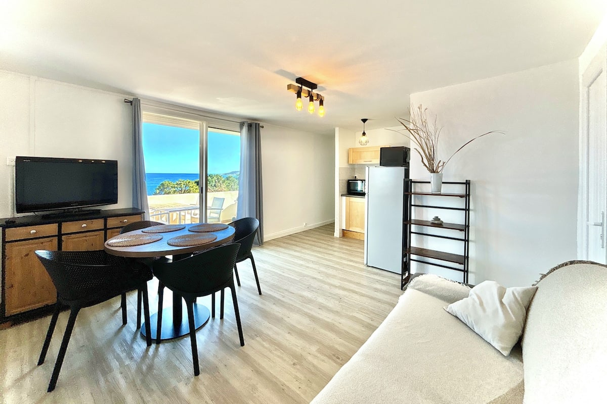 Ein modernes Wohnzimmer mit Fernseher, Esstisch und Stühlen, eine Küche mit Kühlschrank und ein Balkon mit Blick.