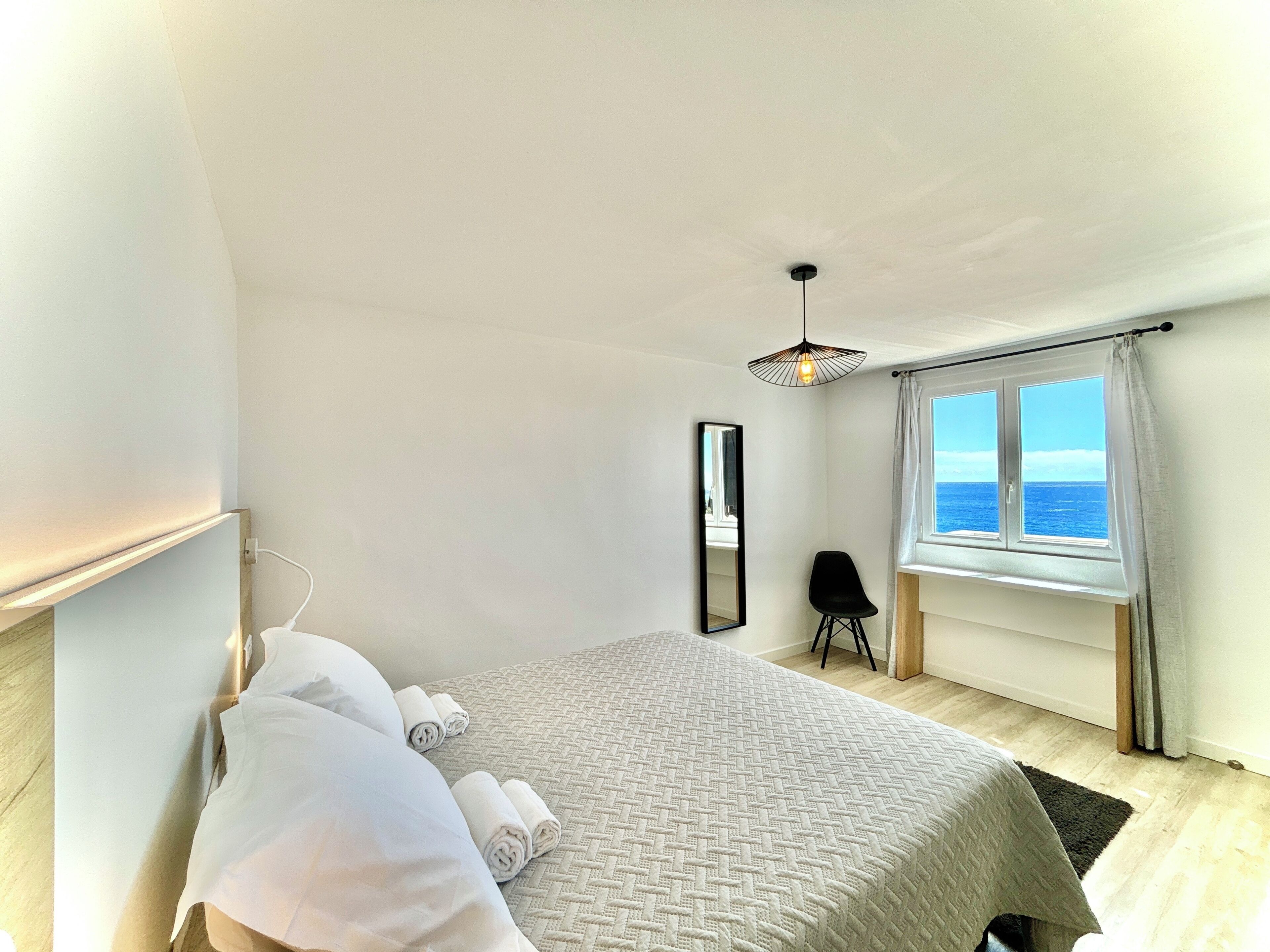Une chambre à coucher avec un lit, une chaise, une fenêtre donnant sur la mer et un luminaire suspendu.