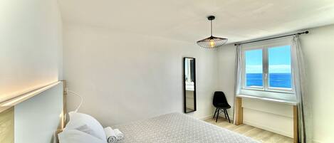 Une chambre à coucher avec un lit, une chaise, une fenêtre donnant sur la mer et un luminaire suspendu.