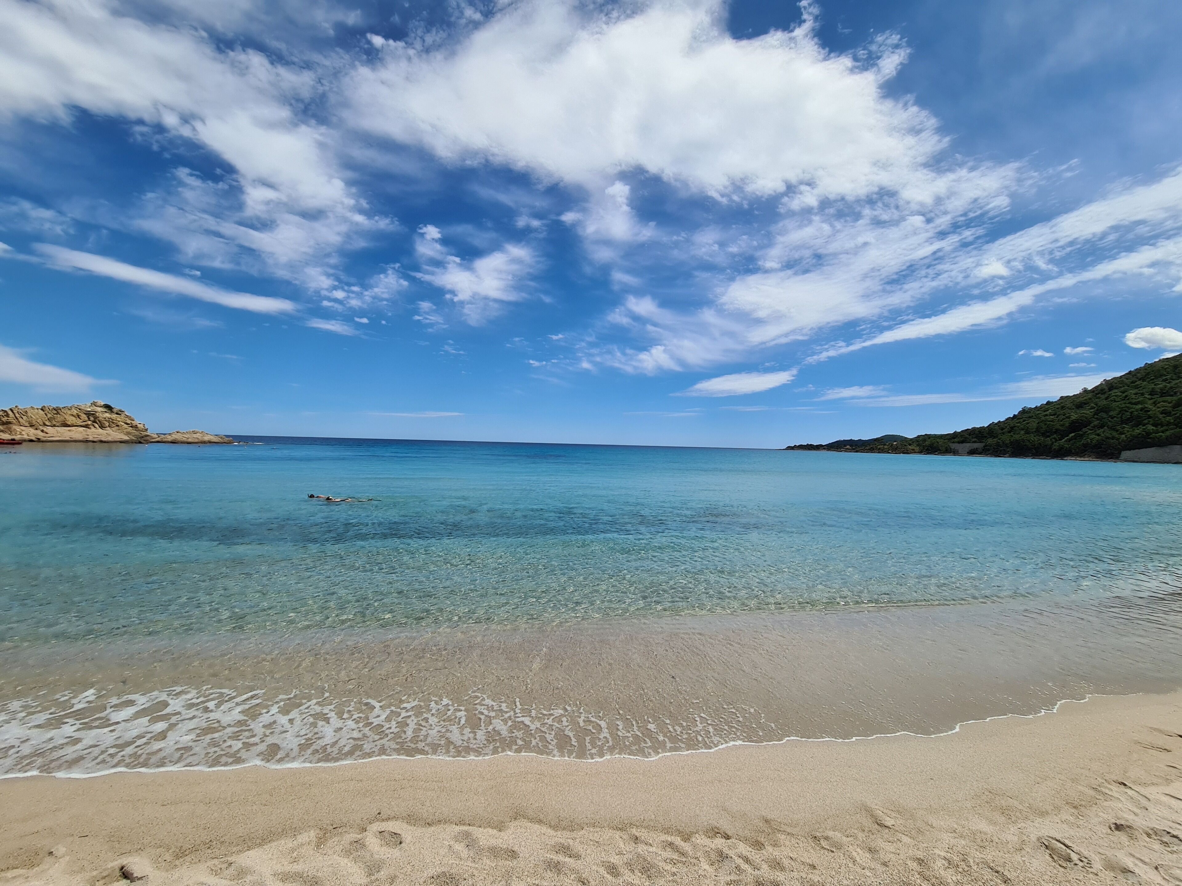 A beach with clear turquoise water, a sandy shore, and a hilly landscape in the background.