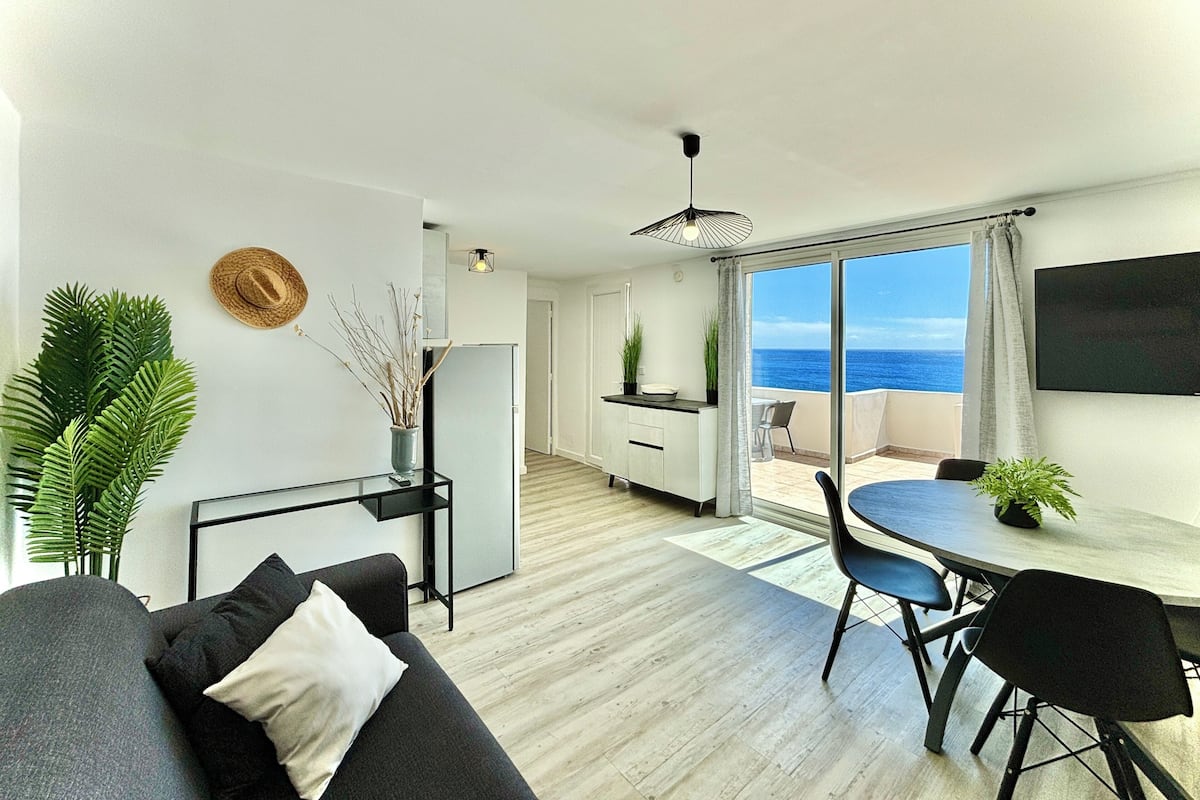 Ein modernes Wohnzimmer mit einer Couch, einem Esstisch und Stühlen. Es gibt eine Küchenzeile mit einem Kühlschrank und einem weiteren Esstisch. Das Zimmer verfügt über große Fenster mit Meerblick.