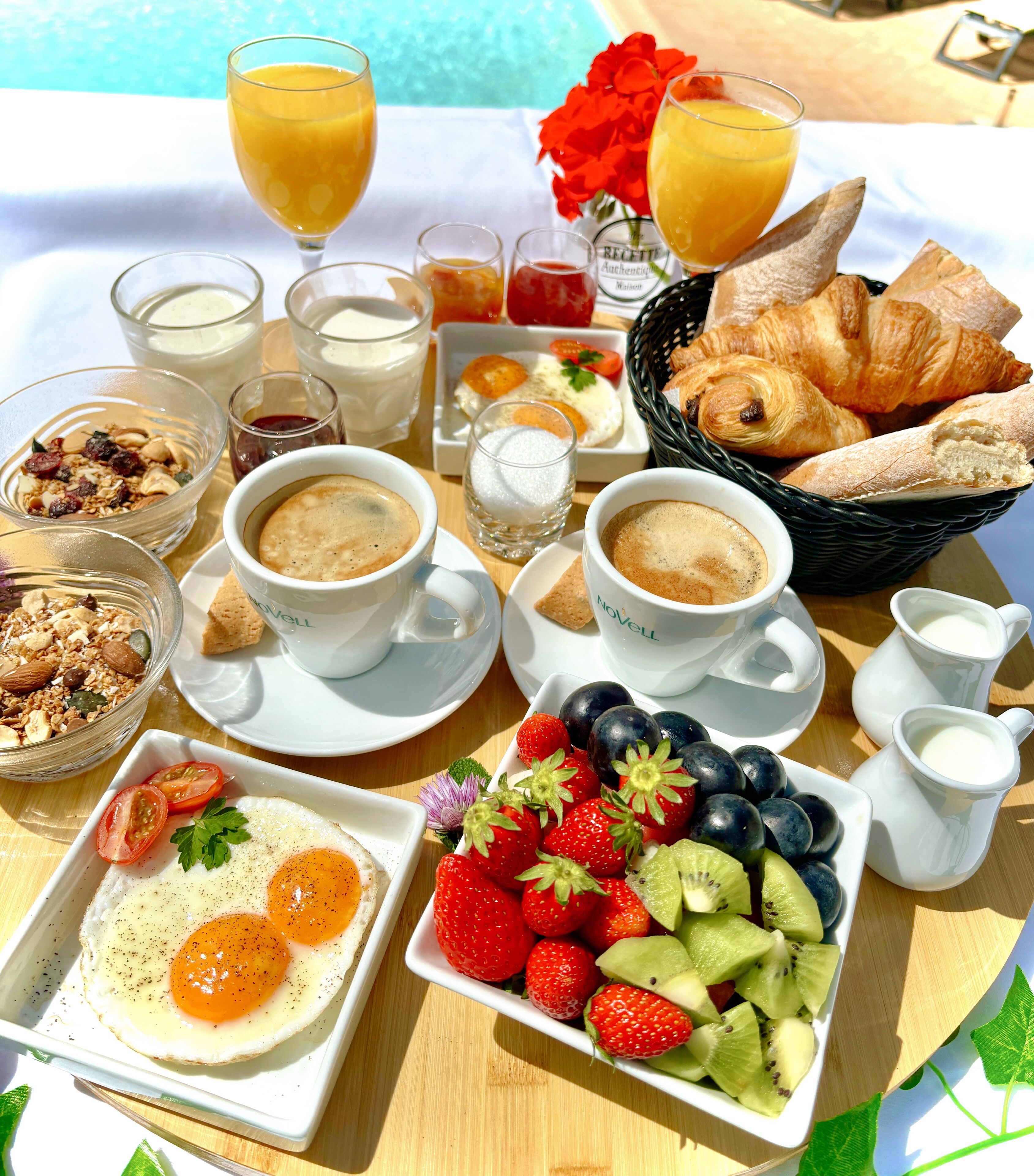 A breakfast spread with croissants, fruit, coffee, and milk on a table by a pool.