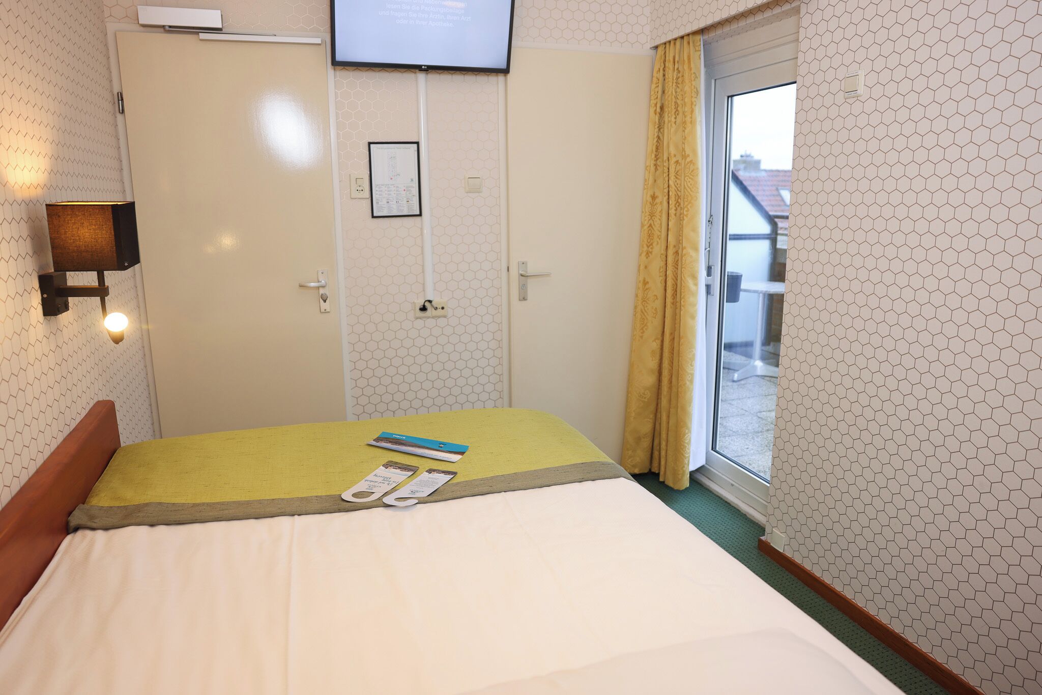 A hotel room with a bed, a television mounted on the wall, a window with curtains, and a door with a calendar.