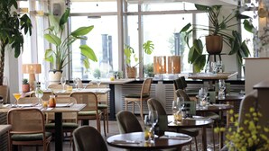 A restaurant with round tables, wicker chairs, and potted plants. - Hotel Noordzee (Katwijk Aan Zee)