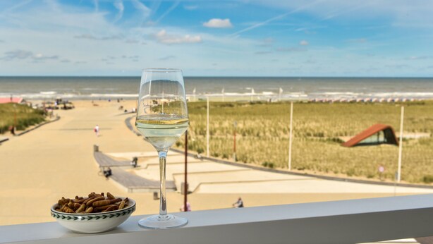 A glass of white wine and a bowl of snacks on a balcony overlooking a beach with a clear sky. - Hotel Noordzee (Katwijk Aan Zee)