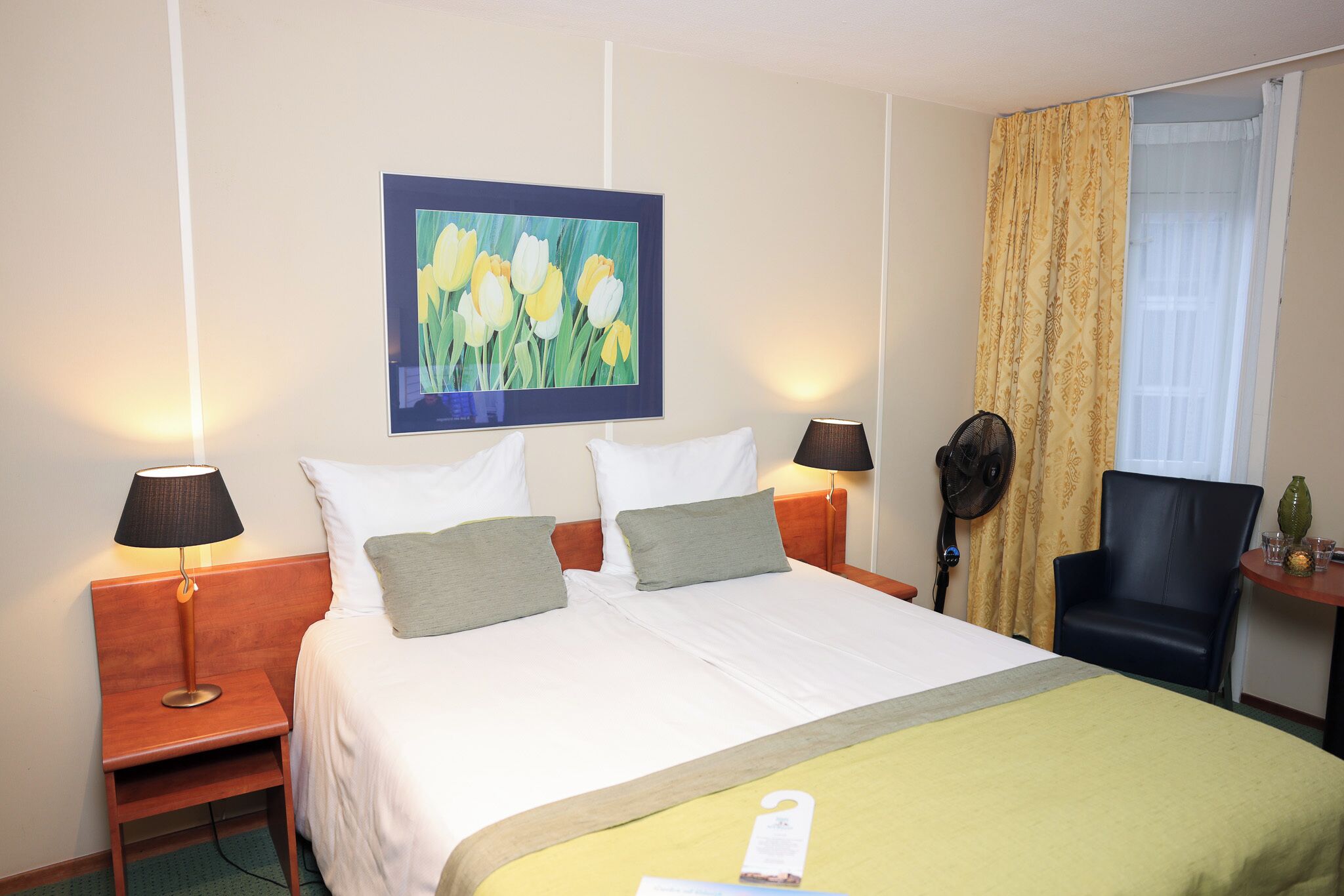 A hotel room with a bed, two bedside lamps, a nightstand, a chair, a table with a vase, and a painting of flowers on the wall.