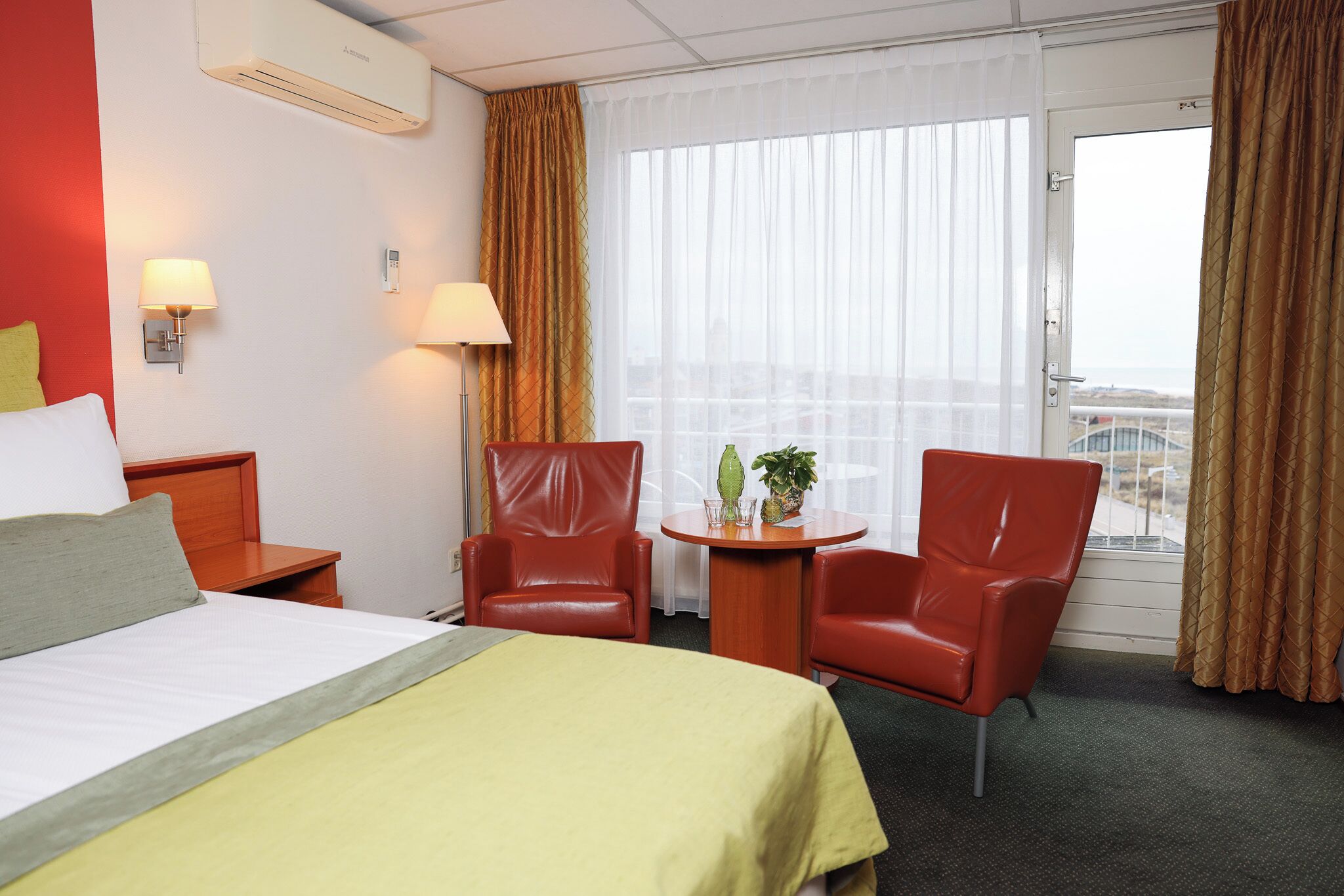 Quarto de hotel com uma cama, duas cadeiras vermelhas, uma mesa pequena com uma planta e uma janela com cortinas.