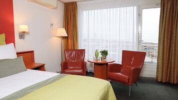 Quarto de hotel com uma cama, duas cadeiras vermelhas, uma mesa pequena com uma planta e uma janela com cortinas.