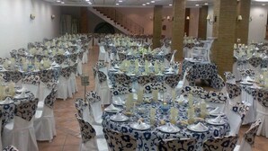 Une salle de banquet avec des tables rondes dressées pour un événement formel, agrémentée de nappes à motifs floraux et de chaises blanches.