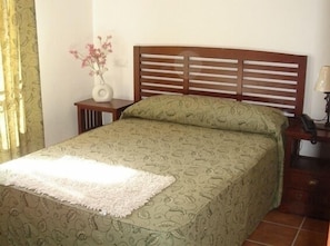 Uma cama com colcha estampada, cabeceira de madeira, mesa de cabeceira com abajur e um vaso com flores.