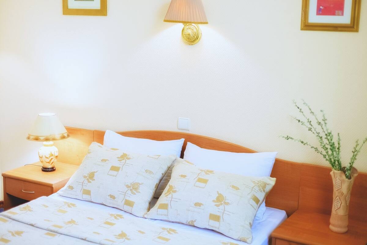 A neatly made bed with floral-patterned pillows, a bedside lamp, and a vase with greenery on a wooden nightstand.