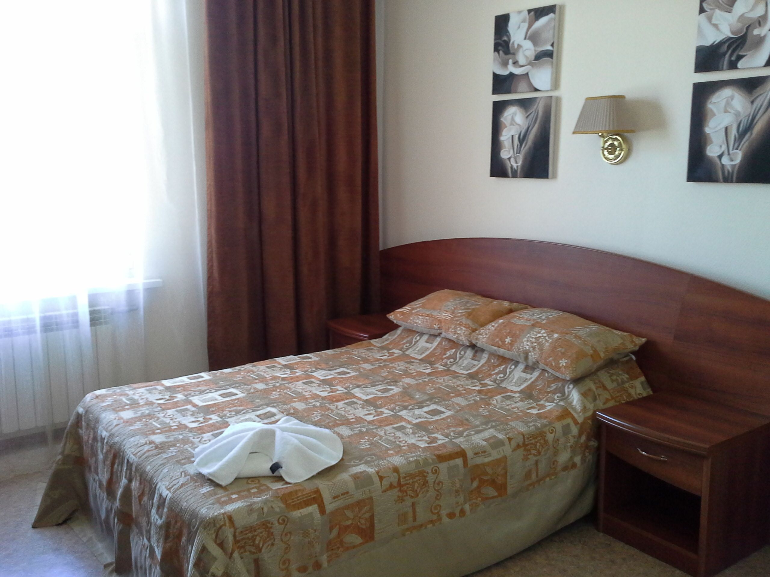 A neatly made bed with a patterned bedspread, a white towel, and two pillows. There is a nightstand with a lamp and a drawer. The room has a window with curtains and artwork on the wall.